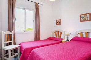 A room with two beds, a chair, a window with curtains, a nightstand with a lamp, and two framed pictures on the wall.