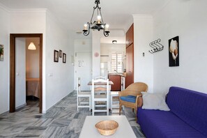 A room with a dining table, chairs, a sofa, a chandelier, and a kitchen area in the background.
