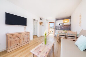 A modern living room with a flat-screen TV, wooden furniture, a dining area, and a kitchen in the background. - Apartment Close to the Beach with Pool Access, Private Terrace, Air Conditioning and Wi-Fi (Canyamel)