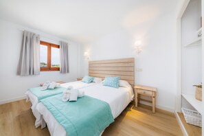 A hotel room with two beds, a wooden headboard, bedside tables, a small bench, and a window with curtains. - Apartment Close to the Beach with Pool Access, Private Terrace, Air Conditioning and Wi-Fi (Canyamel)