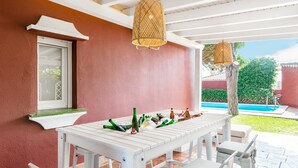 Eine überdachte Terrasse mit einem weißen Tisch und Stühlen, einem hängenden Laternen und einem Pool im Hintergrund.