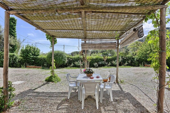 A covered outdoor seating area with a table and chairs, surrounded by gravel and greenery.