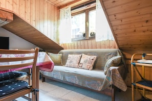 A cozy attic room with a wooden sloped ceiling, a sofa with patterned cushions, a wooden chair, and a small table.