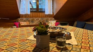 A cozy attic room with a wooden sloped ceiling, a patterned tablecloth, a dining table, a plant, a candle, and a window with curtains. - Apartment “Mansarda tra le Dolomiti di Brenta" with Mountain View & Balcony (Bocenago)
