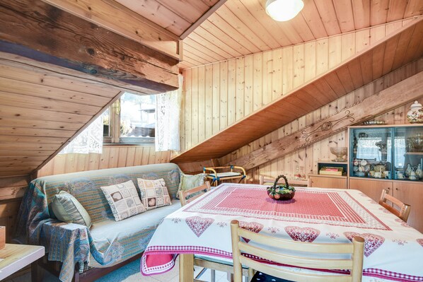 A cozy attic room with wooden walls, a dining table with a red and white tablecloth, a sofa with patterned cushions, and a window with lace curtains.