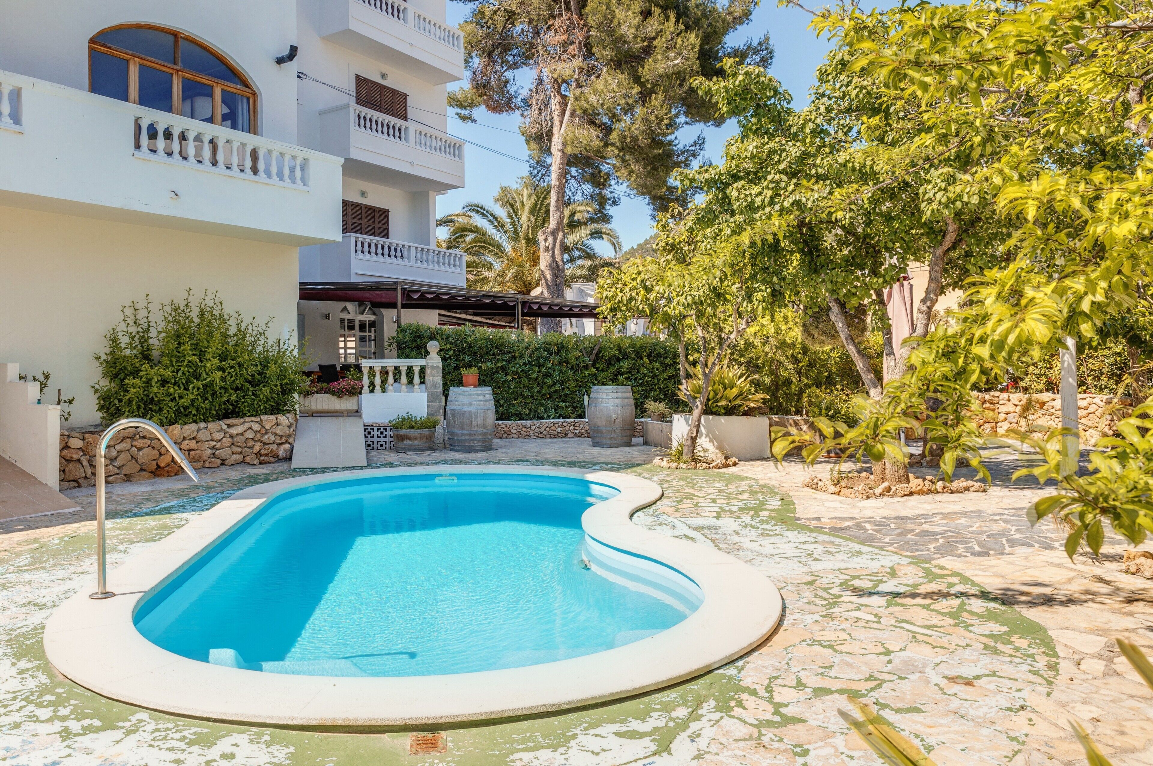 Una piscina rodeada por un patio de piedra, un edificio blanco con balcones y abundante vegetación.