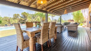 A covered outdoor dining area with a wooden table and wicker chairs, overlooking a pool and greenery.