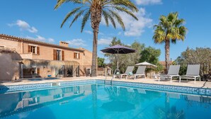 Una piscina con sillones de descanso y sombrillas, rodeada de palmeras y un edificio con techo de tejas.