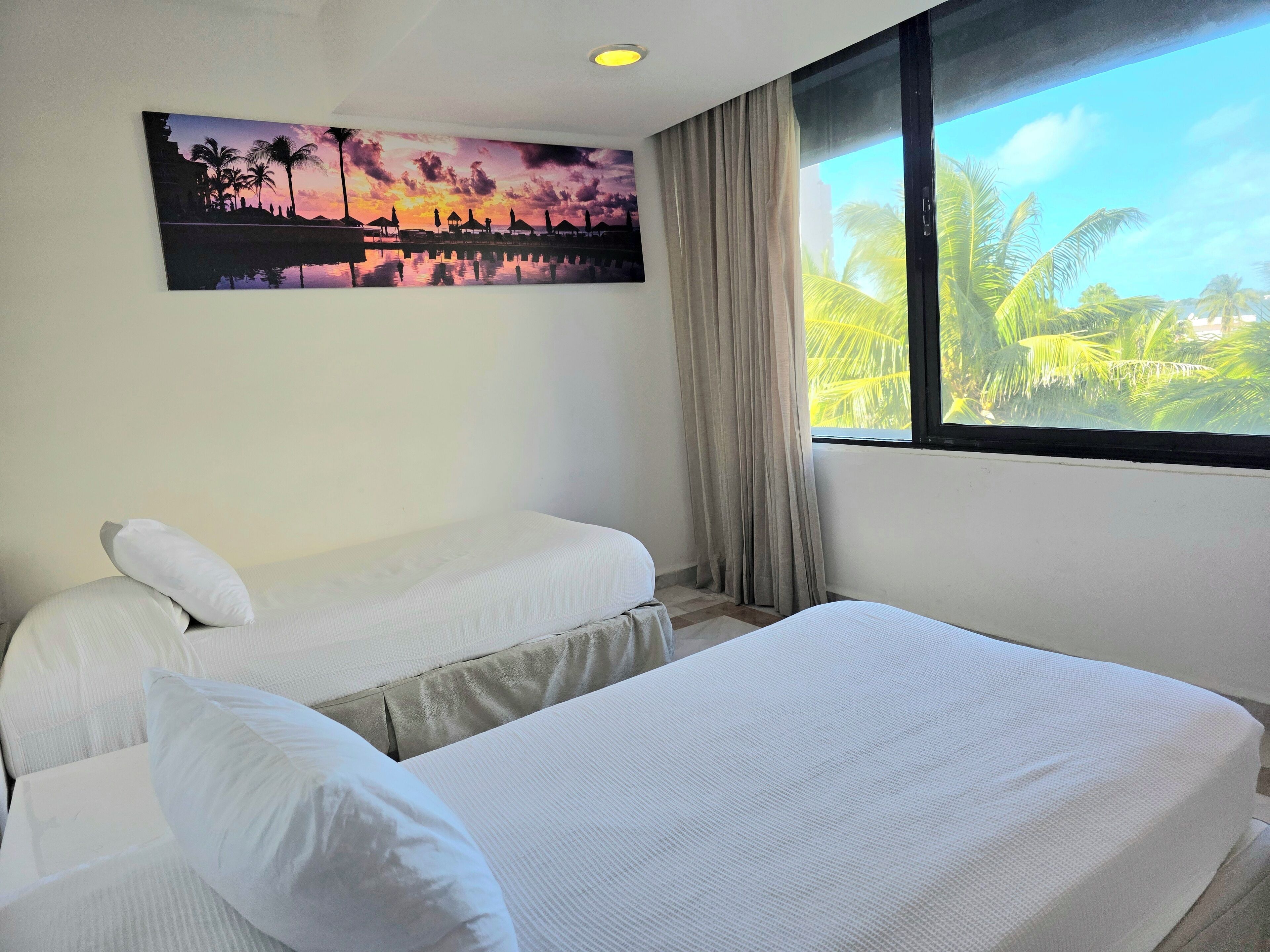 A hotel room with two single beds, a large window with a view of palm trees, and a sunset painting on the wall.