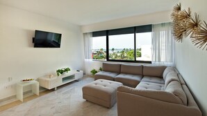 Een moderne woonkamer met een groot raam, een flatscreen televisie, een beige modulaire bank en een klein wit bijzettafeltje met een plant.