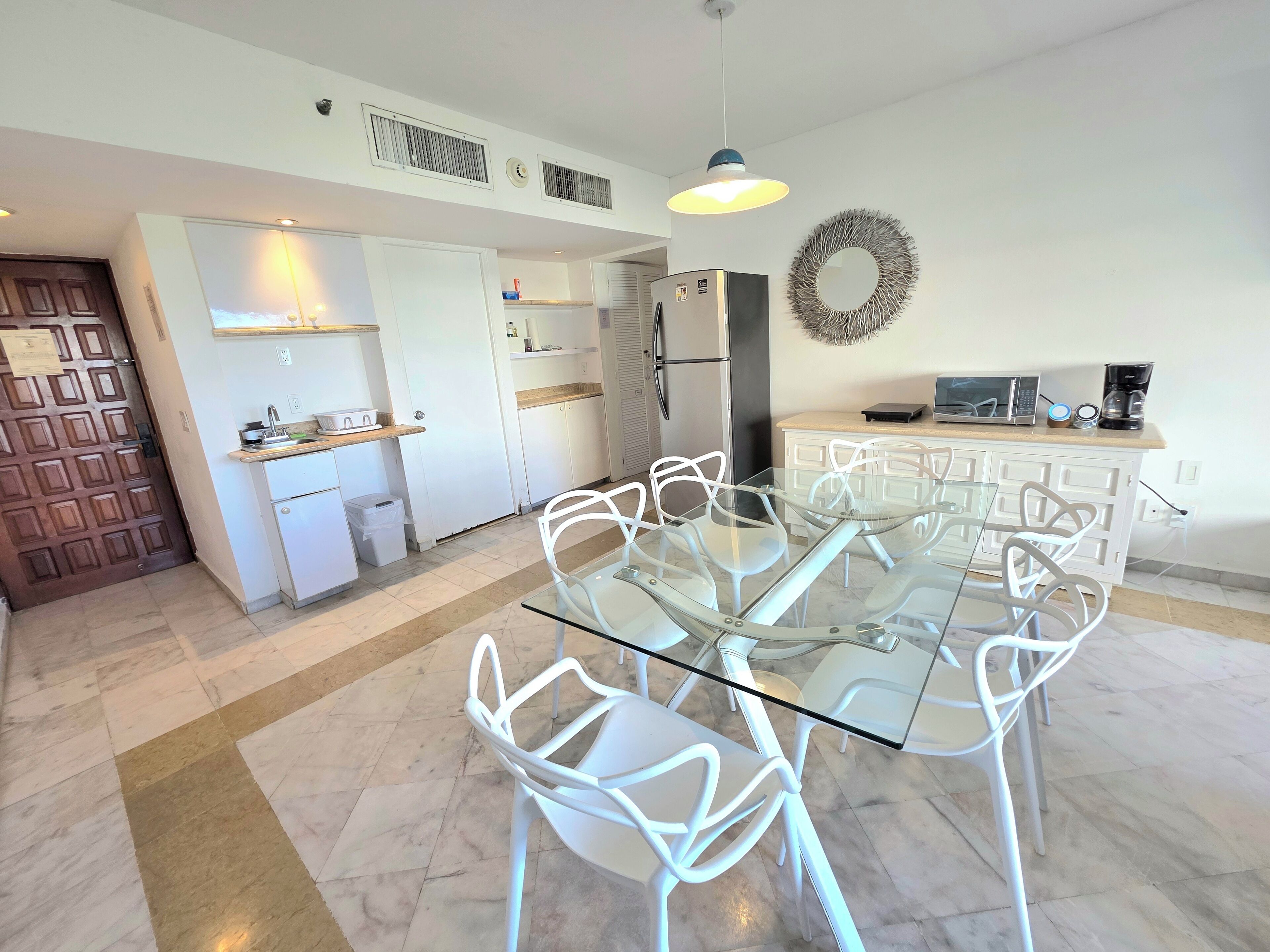 A modern kitchen with white cabinets, a glass dining table, and white chairs. There is a stainless steel refrigerator, a microwave, and a coffee maker on the countertop.