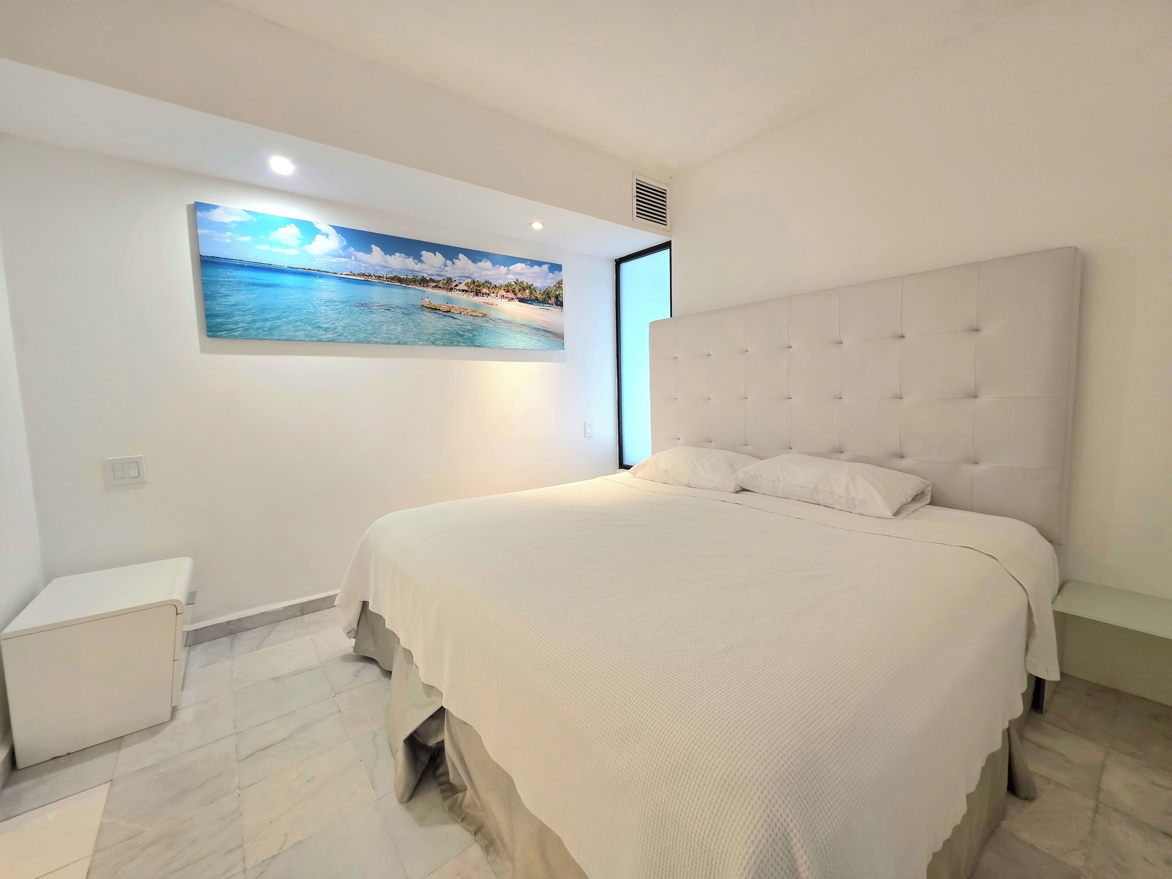 A bedroom with a large bed, a headboard, a nightstand, and a wall-mounted painting of a beach scene.