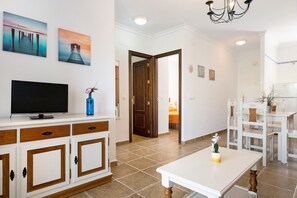 Un salon moderne avec un téléviseur, des meubles en bois, un coin repas avec des chaises blanches et un couloir menant à une autre pièce.
