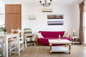 Un salon comprenant une armoire en bois, une table à manger avec des chaises, un canapé recouvert d’un plaid rose, une petite table avec une lampe, et deux tableaux encadrés accrochés au mur.