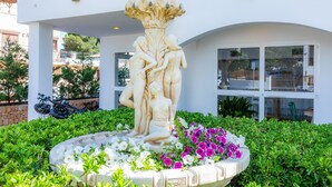 Un bâtiment blanc doté d’un balcon, une fontaine ornée d’une sculpture et un jardin fleuri.