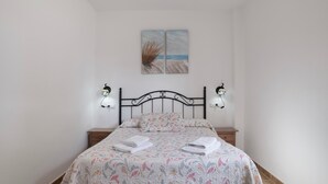 A neatly made bed with a floral bedspread, two wall-mounted lamps, a small bedside table, and a painting of a beach scene above the bed.