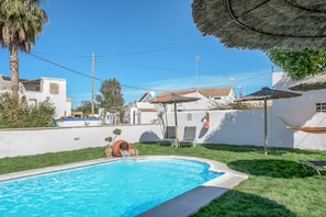 Una piscina con sombrilla de paja, césped y edificios blancos con tejados de tejas rojas.