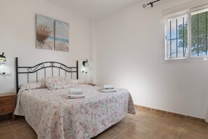 Een slaapkamer met een bed, een nachtkastje, een wandlamp, een raam met tralies en twee schilderijen met strandthema.