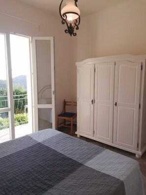 Une chambre à coucher comprenant un lit, une armoire, une chaise et un balcon offrant une vue sur la verdure et les collines.