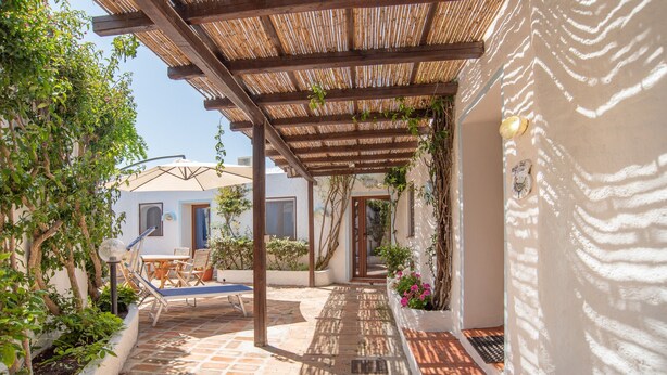 A covered outdoor patio with a pergola, a seating area with a table and chairs, and a pathway leading to a building entrance.
