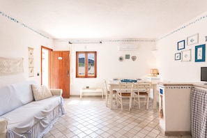 Una habitación con sofá blanco, puerta de madera, mesa de comedor con sillas, una pequeña banca y aire acondicionado instalado en la pared.