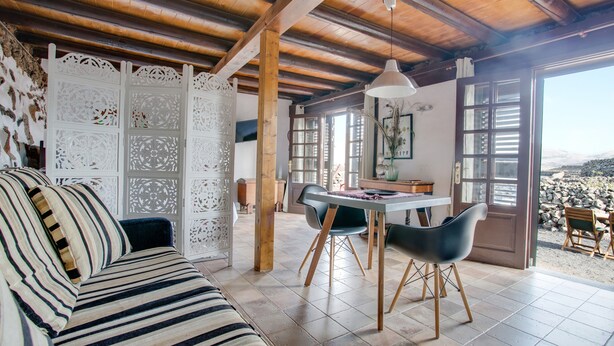 A room with a wooden ceiling, a striped sofa, a dining table with chairs, and a view through a large window. - Rural Stone Cottage "Estudio Pu" with Volcano View, Garden & Wi-Fi (Vegueta Lanzarote)