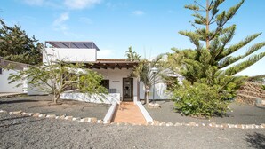 A modern house with a paved driveway, a covered entrance, and a large pine tree in the foreground. - Rural Stone Cottage "Estudio Pu" with Volcano View, Garden & Wi-Fi (Vegueta Lanzarote)