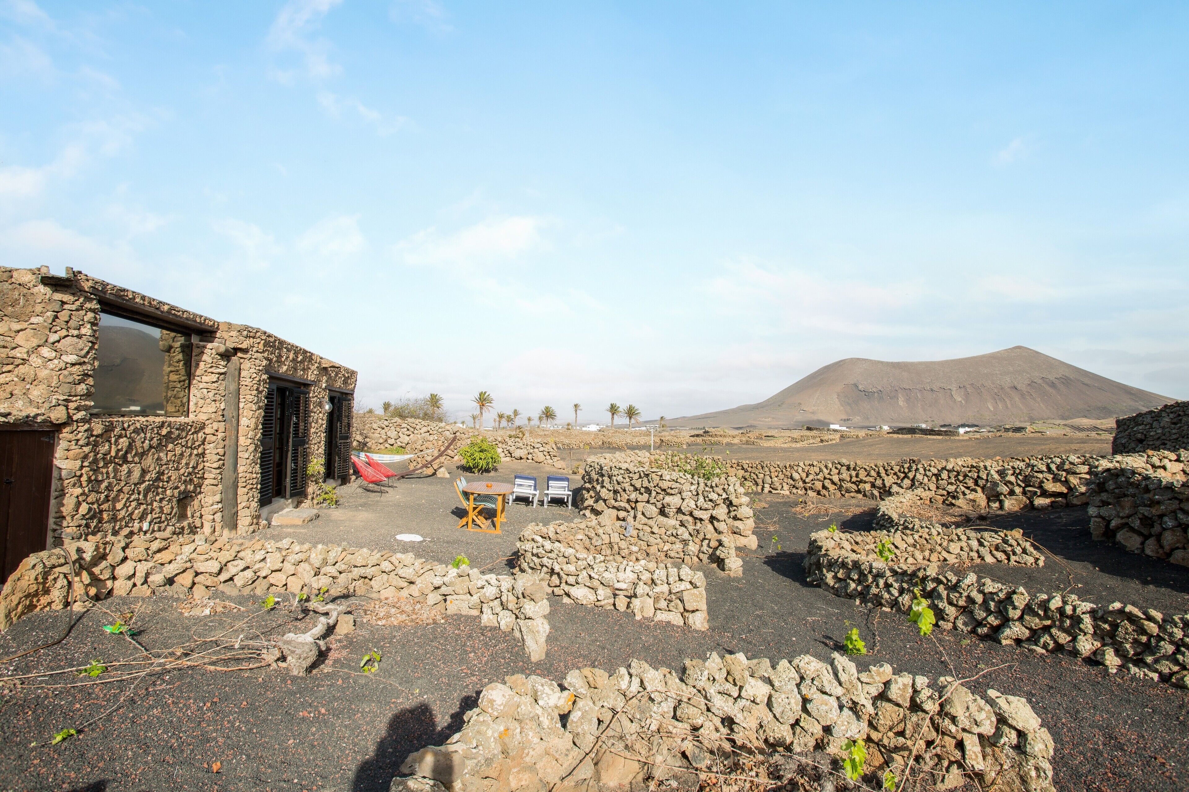 A stone building with a table and chairs outside, surrounded by stone walls and a mountain in the background.