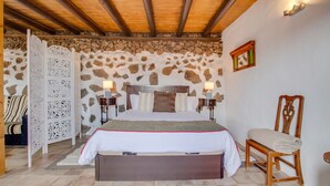 A bedroom with a stone wall, a wooden ceiling, a bed with a headboard, bedside tables, a chair, and a decorative screen. - Rural Stone Cottage "Estudio Pu" with Volcano View, Garden & Wi-Fi (Vegueta Lanzarote)