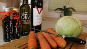 Une planche à découper en bois avec des carottes, un chou et un couteau. On y trouve également des bouteilles d’huile d’olive et une bouteille de vin.