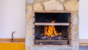 Una chimenea de piedra con fuego encendido en su interior, un estante de madera en la parte inferior y una pared cubierta de azulejos amarillos a la izquierda.