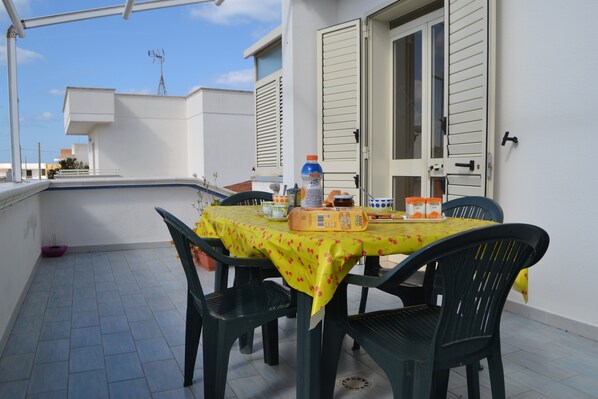 Un balcon avec une table dressée pour deux personnes, des chaises, une bouteille d’eau et une miche de pain.