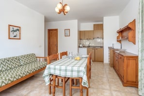 A dining area with a table set for four, a sofa, wooden cabinets, and a kitchen in the background.