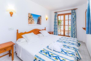 Quarto com cama de madeira, duas mesas de cabeceira, um criado-mudo, janela com cortinas estampadas e luminária fixada na parede.