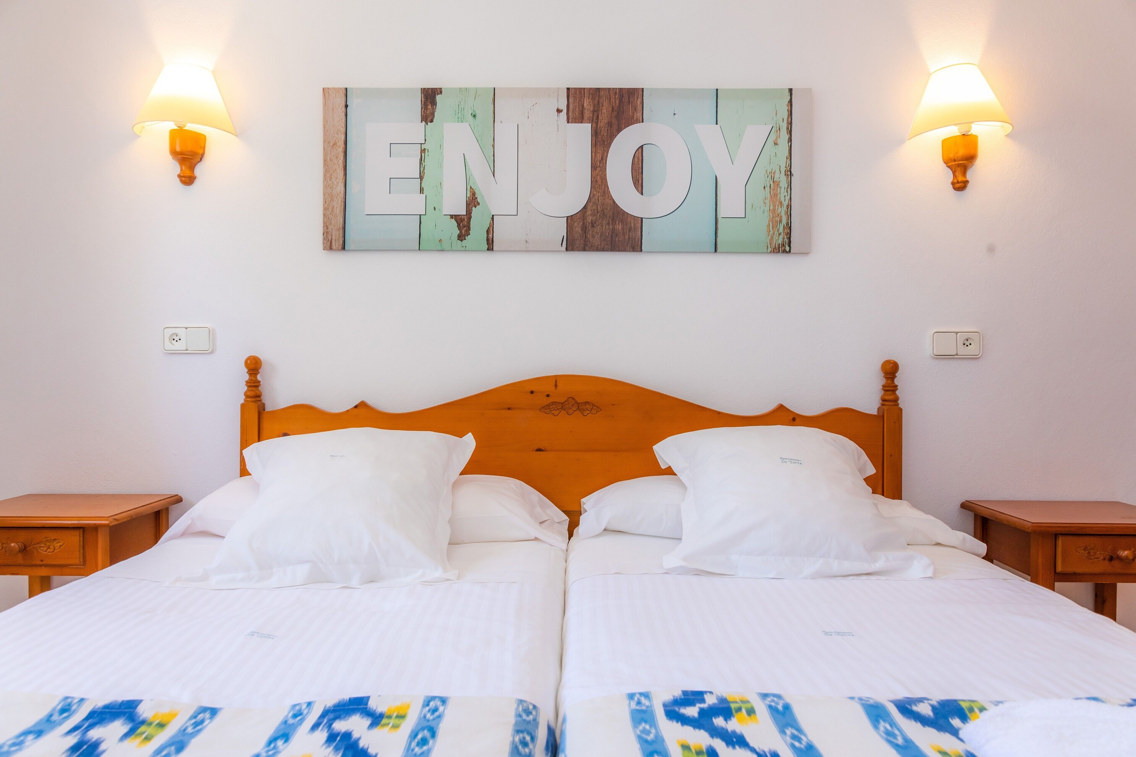 Ein Doppelbett mit weißen Leinen, zwei Nachttische, ein Holzkopfstück und ein an der Wand befestigtes Schild mit der Aufschrift 'ENJOY'.