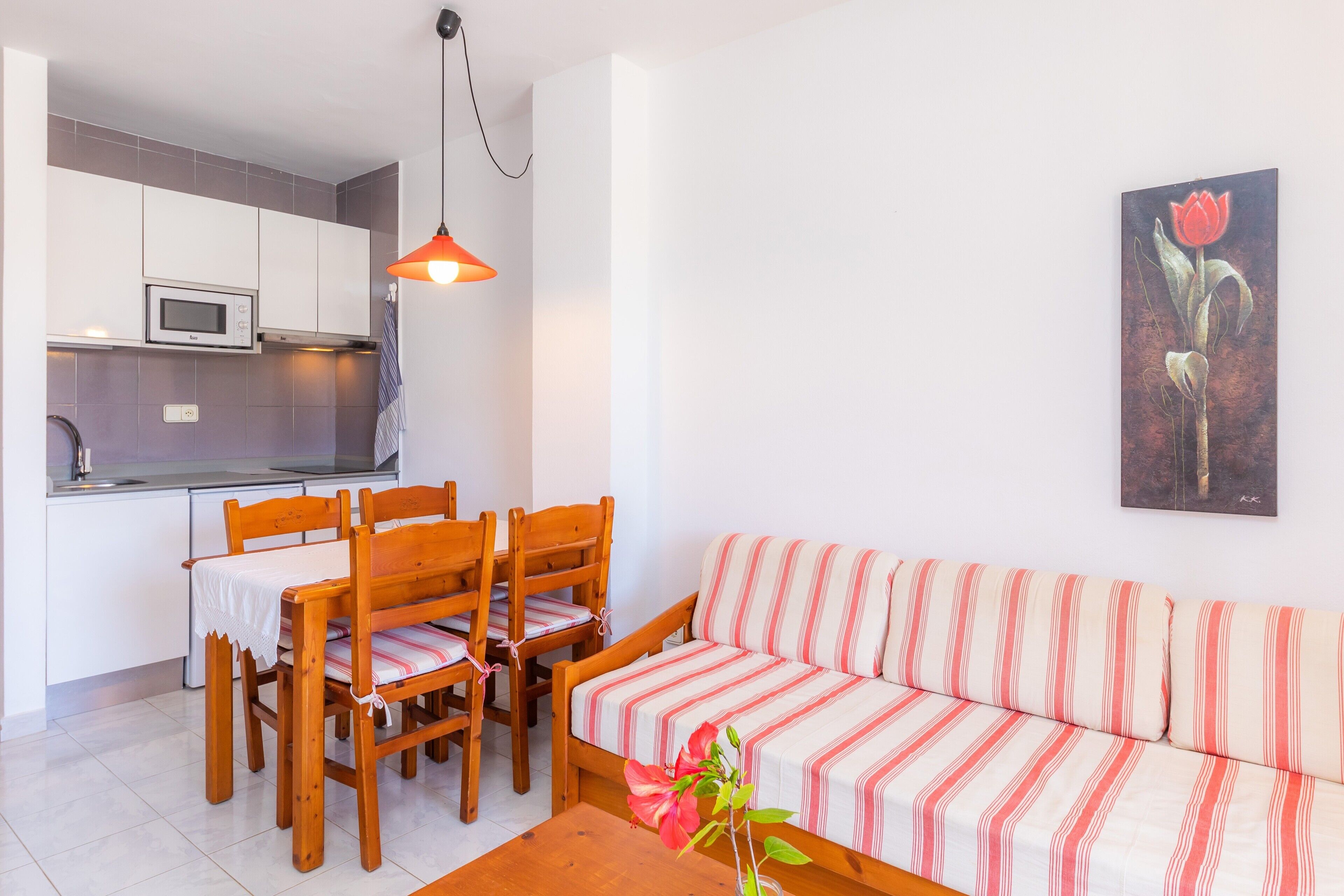 Une cuisine moderne avec des meubles blancs et un four à micro-ondes, un coin repas avec des chaises en bois et une table, et un canapé à rayures agrémenté d’un vase de fleurs.