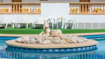 Une piscine agrémentée d’une sculpture en pierre, de chaises longues blanches et d’une pelouse verdoyante.