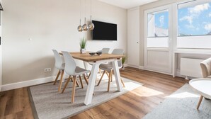 Un comedor con una mesa de madera, sillas blancas, un televisor montado en la pared y un ventanal grande.