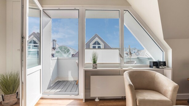 Una habitación moderna con techo inclinado, un radiador blanco, un sillón beige, una planta pequeña en maceta y vistas a casas vecinas a través de grandes ventanales.