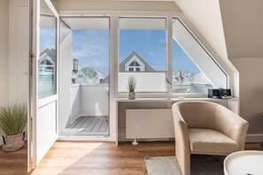 A modern room with a sloped ceiling, a white radiator, a beige armchair, a small potted plant, and a view of neighboring houses through large windows.