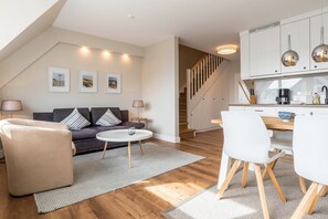 A modern living room with a sofa, coffee table, and chairs, a kitchen with white cabinets and a dining area.