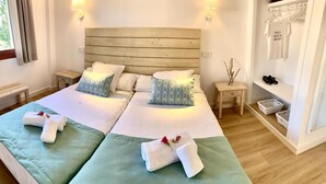 Une chambre double avec deux lits, des tables de chevet, une petite table, une fenêtre avec des rideaux, une lampe fixée au mur, un porte-chaussures et une petite étagère avec des paniers.