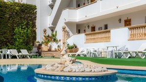 Une piscine avec des chaises longues, une fontaine décorative et un bâtiment doté de balcons.