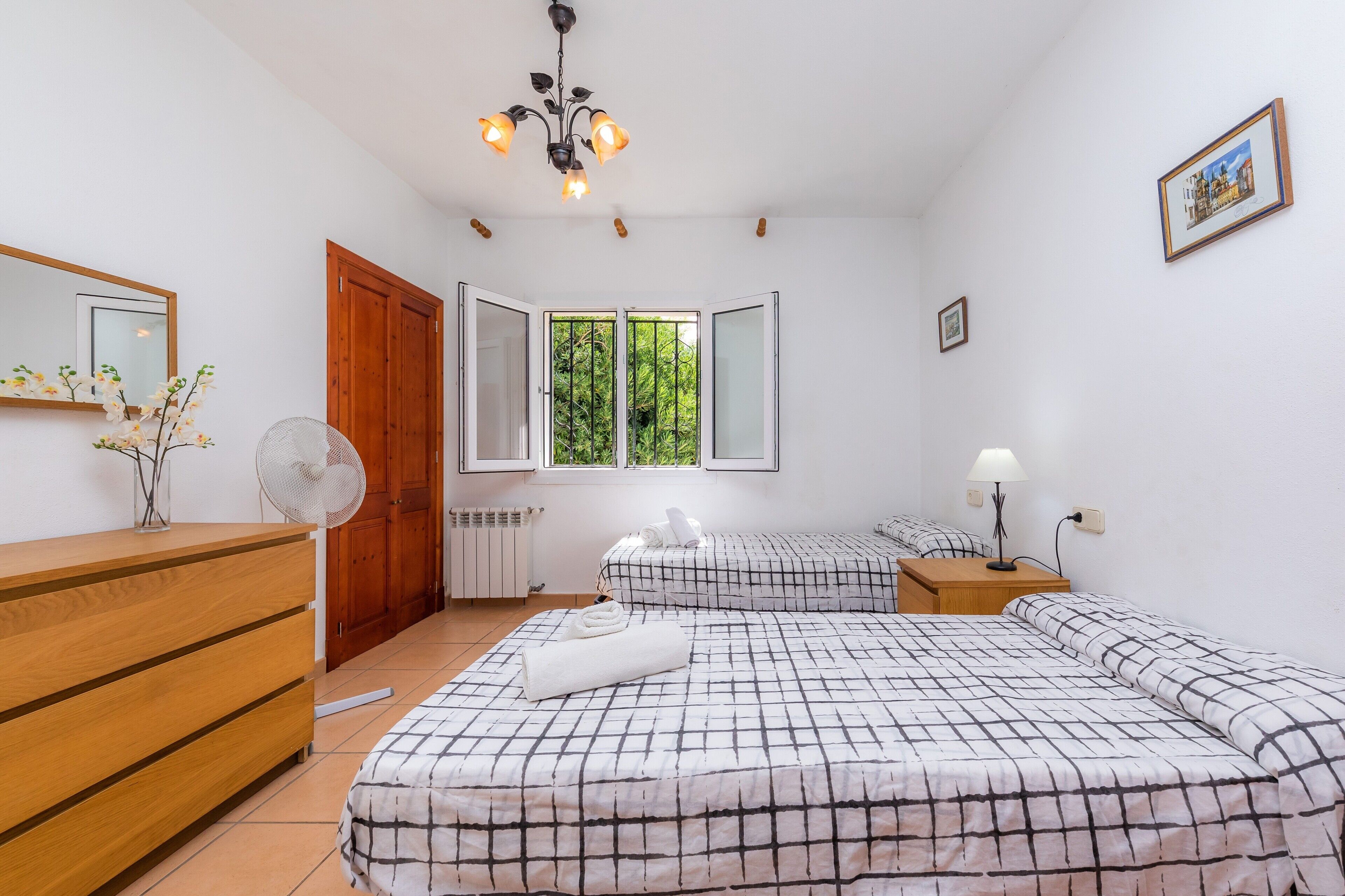 Een slaapkamer met twee bedden, een houten dressoir, een spiegel, een ventilator, een radiator, een raam met uitzicht op groen en een kroonluchter.