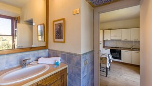Baño con lavamanos, espejo y perchero. Hay una puerta que da a una cocina con armarios y estufa.