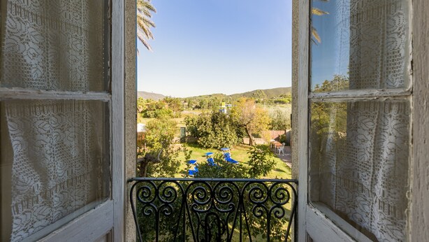 Vista a través de una ventana con cortinas de encaje, un balcón con barandilla de hierro forjado y un paisaje con árboles y colinas.