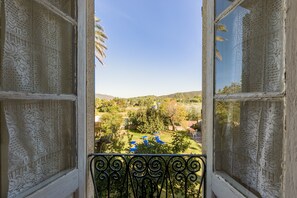 Vista a través de una ventana con cortinas de encaje, un balcón con barandilla de hierro forjado y un paisaje con árboles y colinas.