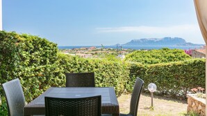 Eine Terrassengruppe im Freien mit Tisch und Stühlen, freier Blick auf das Meer und ferne Berge, und ein gepflegter Heckenstreifen.