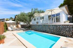 Une piscine bordée d’un muret en pierre, avec des chaises longues blanches et des parasols.