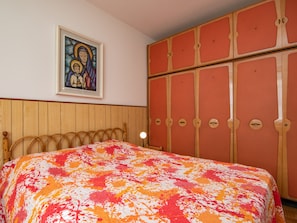 A bedroom with a wooden headboard, a bed with a colorful blanket, a framed painting on the wall, and a large red wardrobe.